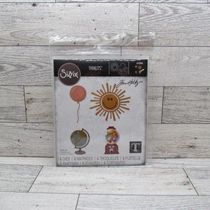 May include: A package of Sizzix Thinlits dies featuring various designs, including a smiling sun, a balloon, a globe, and a gumball machine. The package includes the text "6 DIES" and "Jim Holtz".