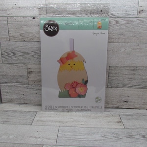 Puede incluir: Un juego de troqueles Sizzix Thinlits con un pollito de Pascua troquelado que emerge de una cáscara de huevo agrietada, adornado con un lazo rosa y flores. El embalaje incluye el texto "12 DIES" y "Georgia Eung".