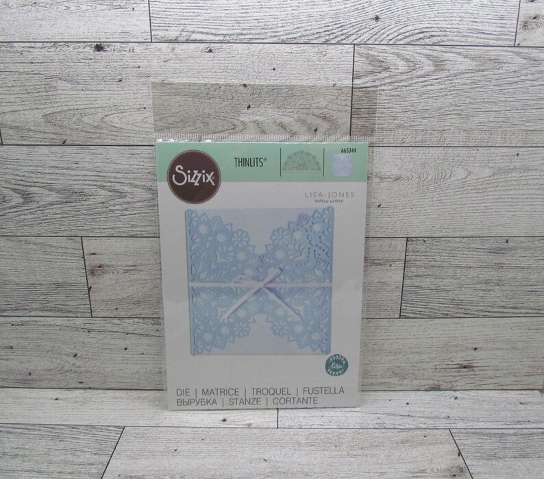 May include: A Sizzix Thinlits die set with a light blue, floral design. The package includes the text "LISA JONES" and die cutting terms in multiple languages. The die-cut design is tied with a white ribbon.