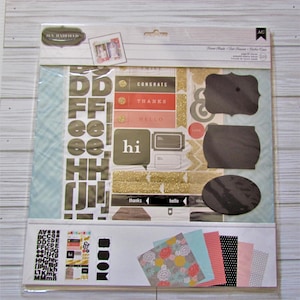 May include: A scrapbook kit by Jen Hadfield, featuring various embellishments. Includes letter stickers, decorative labels with phrases like "Congrats" and "Thanks," and patterned paper in floral and geometric designs. The kit is packaged in a clear plastic sleeve.