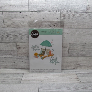 May include: Sizzix Thinlits die set with a design of two owls under an umbrella, and the phrases "just us two" and "always love you." The set contains 9 dies for papercrafting. The package is sealed in clear plastic.