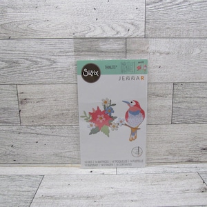 Puede incluir: Un paquete de troqueles Sizzix Thinlits con un pájaro posado en una rama con flores. El pájaro tiene colores rojo, rosa y azul. El paquete incluye 14 troqueles y el nombre "JENNAR".
