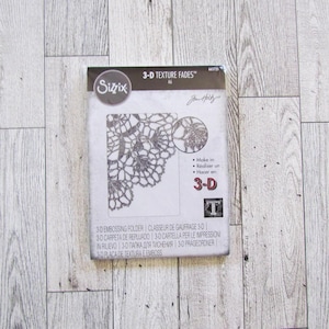 May include: A Sizzix 3-D Texture Fades embossing folder package. The package features a detailed floral lace design. The package also includes the text "3-D" and the Sizzix logo.