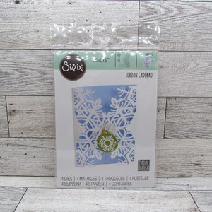 May include: A Sizzix die set with a snowflake design. The package shows a card with a white snowflake cut-out, a green and white ornament, and the text "JORDAN CADERAO". The package also includes the text "4 DIES | 4 MATRICES | 4 TROQUELES | 4 FUSTELLE".