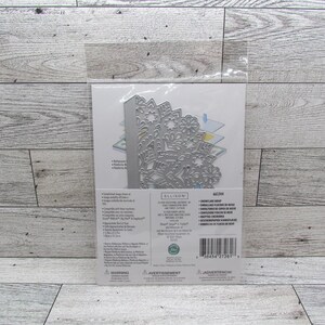 May include: A silver metal die cut in a clear plastic package. The die cut features a detailed snowflake design. The package includes text and is on a light wood grain surface.
