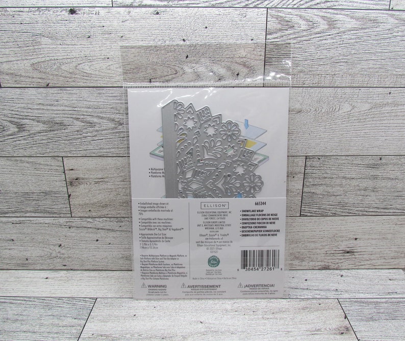 May include: A silver metal die cut in a clear plastic package. The die cut features a detailed snowflake design. The package includes text and is on a light wood grain surface.