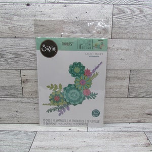 May include: A Sizzix Thinlits die set by Lisa Jones, featuring floral designs in shades of green, pink, and purple. The set includes 15 dies for creating paper flowers and foliage. The package is sealed in clear plastic.