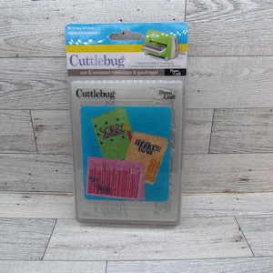 May include: A Cuttlebug die-cutting and embossing machine kit with a blue and white package. The kit includes a cutting die and embossing folder. The kit includes a blue felt square with three small cards that say "Sorry I missed it", "It's your day", and "Make a wish".