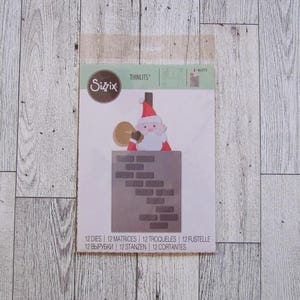Puede incluir: Juego de troqueles Sizzix Thinlits con un diseño de Papá Noel. La imagen muestra una figura de Papá Noel saliendo de una chimenea de ladrillo. El embalaje incluye el texto "12 DIES | 12 MATRICES | 12 TROQUELES | 12 FUSTELLE | 12 ВЫРУБКИ | 12 STANZEN | 12 CORTANTES".