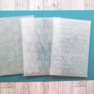 May include: Three rectangular plastic embossing folders with translucent surfaces. The left folder features various Christmas ornaments, the center folder displays a pattern of pine trees, and the right folder contains holiday text including Merry Christmas.