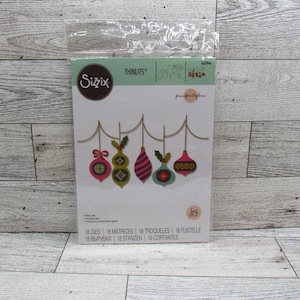 May include: A Sizzix Thinlits die set featuring six festive Christmas ornament designs in pink, green, and gold. The ornaments hang from a gold line. The package includes 18 dies and 18 matrices. The text on the package reads '18 Dies'.
