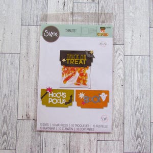 Puede incluir: Un juego de troqueles Sizzix Thinlits con diseños de Halloween. El juego incluye troqueles para una bolsa de caramelos con el texto "Trick or Treat", y pancartas con los textos "Hocus Pocus" y "Boo!", además de un fantasma y un murciélago. El paquete incluye 10 troqueles.