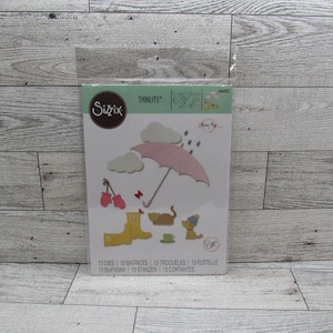 May include: A Sizzix Thinlits die set with a weather theme, including a pink umbrella, rain, clouds, boots, a cat, and a dog. The package contains 13 dies. The text on the package includes the word 'Thinlits'.