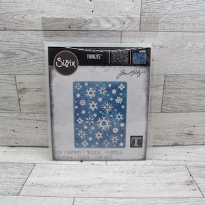 May include: A Sizzix Thinlits die set with a snowflake pattern. The design is a square of blue with white snowflake shapes. The package includes the designer's signature and the word "Thinlits".