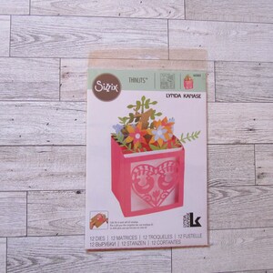 May include: A Sizzix Thinlits die set by Lynda Kanase, featuring a pink box design with a heart-shaped cutout and floral embellishments. The package includes 12 dies for paper crafting. The box design is filled with colorful paper flowers and greenery.