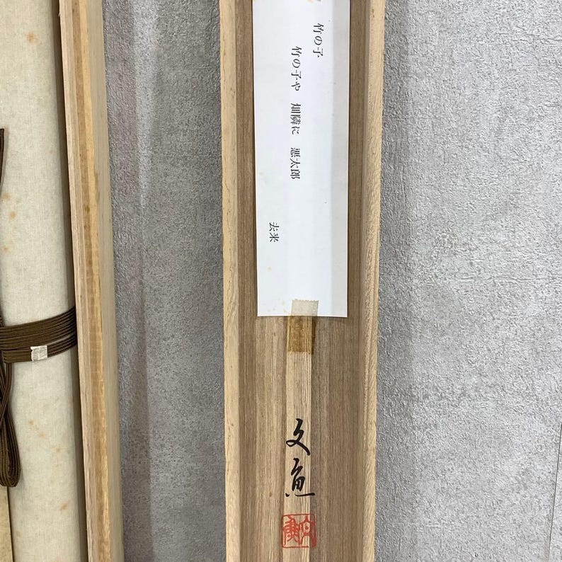 May include: A wooden box with a scroll inside. The box is light brown with a paper label that has Japanese text. The scroll is wrapped with a dark brown cord. The box has Japanese characters on the bottom.