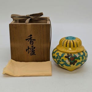 May include: A yellow ceramic incense burner with a turquoise top and floral designs. It is accompanied by a wooden box with Japanese characters and a beige cloth. The burner has a bulbous shape and intricate patterns.