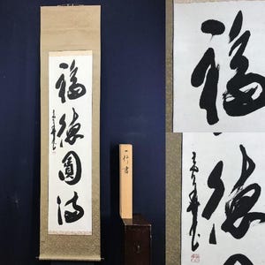 May include: A traditional Japanese calligraphy scroll featuring bold black ink characters on white paper. The scroll is mounted on a tan backing and includes a wooden storage box. The calligraphy is a form of art.