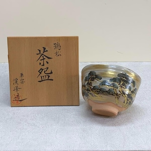 May include: A Japanese tea bowl with a landscape design. The bowl has a gold rim and a painted scene of trees in green and gold. It is presented with a wooden box with Japanese characters.