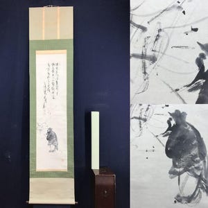 May include: A traditional Japanese scroll painting with calligraphy and a figure in ink wash. The scroll features a green border and a light-colored top and bottom. A small, light green rectangular object sits next to the scroll.