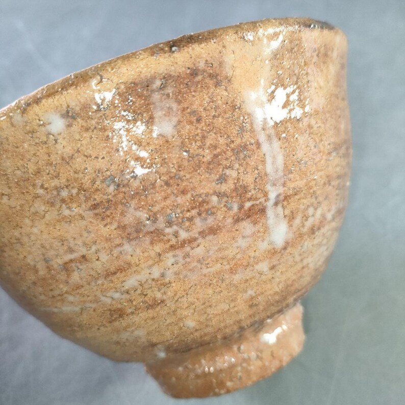 May include: A handmade ceramic bowl with a textured, earthy appearance. The bowl features a warm, brown color with lighter, white streaks and a small, rounded base. The interior is visible, showcasing the bowl's depth and shape.