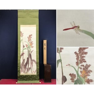 Op de afbeelding: Een antiek Japans rolschilderij met een aquarel van een libelle en bloeiende planten. De rol wordt getoond met een houten steun en een donkerbruine doos. Het kunstwerk heeft een groene rand.
