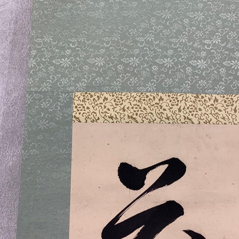 May include: A traditional Japanese scroll with calligraphy. The scroll features a light pink background with bold black brushstrokes. The top border has a floral pattern in shades of gold and brown, and the top is a light green floral pattern.