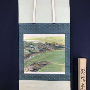 Op de afbeelding: Een traditioneel Japans rolschilderij met een landschap met bergen, een groen veld en water. De rol heeft een lichtgroene achtergrond, een donkerblauwe patroonrand en witte ophanglinten. Een houten label met Japanse karakters staat rechts.