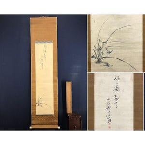 May include: A vintage Japanese scroll painting featuring a minimalist ink wash illustration of an orchid. The scroll is encased in a brown paper with a wooden roller at the bottom. The painting includes Japanese calligraphy.