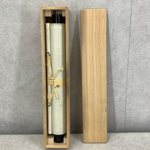Puede incluir: Un rollo enrollado en una caja de madera. El rollo est&aacute; atado con un cord&oacute;n beige. La caja es rectangular y de madera clara. El rollo es blanquecino con detalles en negro. La caja est&aacute; abierta, revelando el rollo y su contenido.