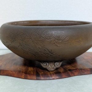 Könnte beinhalten: Eine braune, runde Keramikschale mit geätzten botanischen Designs, die auf einem Holzsockel steht. Die Schale hat einen verzierten Sockel. Der Holzsockel hat eine einzigartige Form. Die Schale dient wahrscheinlich dekorativen Zwecken.