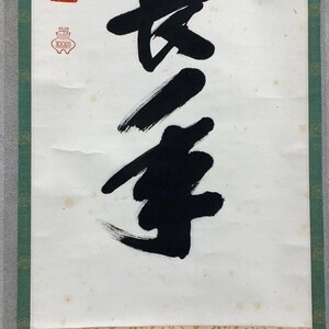 Puede incluir: Un rollo de caligraf&iacute;a japonesa tradicional con audaces pinceladas negras sobre papel blanco envejecido. La obra de arte incluye un sello rojo y un borde verde, mostrando una est&eacute;tica cl&aacute;sica. La caligraf&iacute;a es el punto focal.