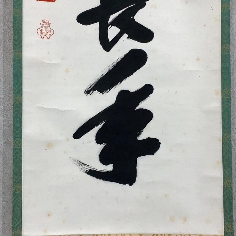 Puede incluir: Un rollo de caligraf&iacute;a japonesa tradicional con audaces pinceladas negras sobre papel blanco envejecido. La obra de arte incluye un sello rojo y un borde verde, mostrando una est&eacute;tica cl&aacute;sica. La caligraf&iacute;a es el punto focal.