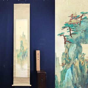 May include: A long, vertical scroll painting depicting a mountain landscape with a waterfall. The artwork has a beige background with a detailed depiction of mountains, trees, and buildings in shades of green, blue, and brown. A wooden box sits to the right.