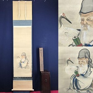 May include: A Japanese scroll painting depicting an elderly man with a long white beard, a crane, and a scroll. The artwork uses muted tones of beige, blue, and grey, with a wooden box on the side.