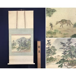 May include: An antique Japanese scroll painting depicting a landscape with mountains, trees, and figures. The scroll is cream-coloured with a light blue border and a wooden hanging rod. The painting shows a man leading a horse.