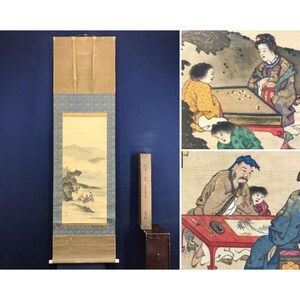 May include: An antique Japanese scroll painting with a landscape scene, displayed with a wooden box and a separate artwork depicting figures playing a board game and painting. The scroll features a mountain and river scene.