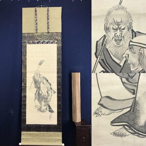 Könnte beinhalten: Traditionelle japanische Rollbildmalerei mit einer detaillierten Tuschezeichnung einer Figur mit Stab. Das Kunstwerk ist mit dekorativen Bordüren gerahmt und auf einem cremefarbenen Hintergrund montiert. Die Schriftrolle wird vor einem dunkelblauen Hintergrund präsentiert.