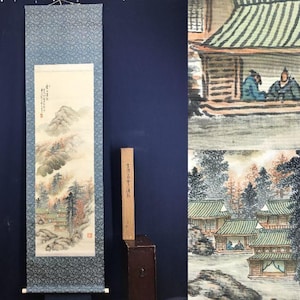 Op de afbeelding: Een antiek Japans rolschilderij dat een landschap met bergen, bomen en traditionele gebouwen afbeeldt. Het kunstwerk heeft gedempte kleuren en is omlijst door een blauwe patroonrand. Een houten kist met Japanse tekst staat naast de rol.