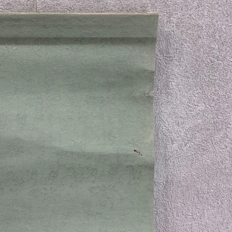 May include: A section of a pale green scroll with a subtle floral pattern. The scroll is partially unrolled, revealing a textured surface. The right side of the image shows a textured, light gray surface.