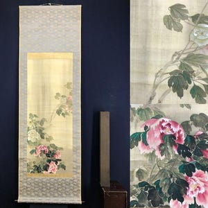 May include: A traditional Japanese scroll painting featuring a floral design. The artwork showcases pink and white peonies with green leaves against a light yellow background. The scroll has a light blue patterned border and a gold accent.
