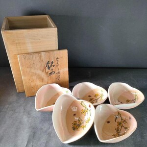 Könnte beinhalten: Fünf herzförmige Keramikschalen mit floralen Mustern in Rosa, Pfirsich und Gelb. Dazu eine Holzkiste und ein Deckel mit japanischer Kalligraphie. Ideal zum Servieren.