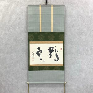 May include: A traditional Japanese scroll painting with calligraphy in black ink on a white background. The scroll features a green and gold patterned border and a light green fabric top and bottom. The scroll is hanging on a wall.