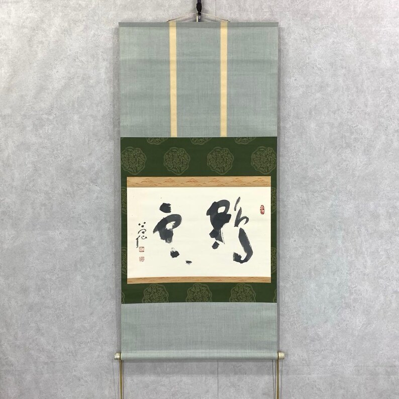 May include: A traditional Japanese scroll painting with calligraphy in black ink on a white background. The scroll features a green and gold patterned border and a light green fabric top and bottom. The scroll is hanging on a wall.