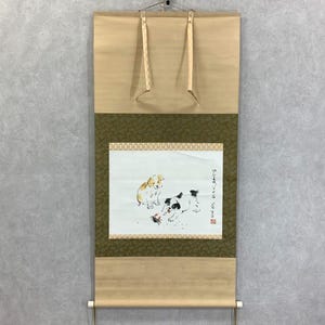 May include: A traditional Japanese scroll painting featuring two puppies. One puppy is tan, and the other is black and white. The painting is on a white background with a green border and tan scroll.