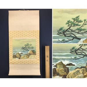 May include: A Japanese scroll painting depicting a coastal scene with a windswept tree and crashing waves. The artwork features a square central image with a patterned border, and is mounted on a cream-colored backing.