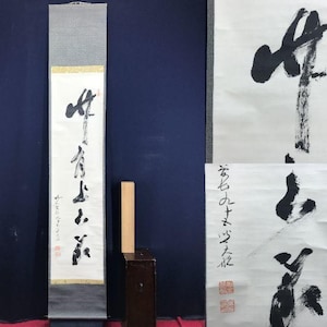 May include: A long, vertical Japanese scroll with black calligraphy on white paper. The scroll is framed by a gray border and a gold accent. A small wooden block and a dark brown cabinet are next to the scroll.