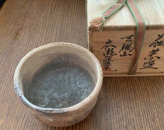 Könnte beinhalten: Eine Keramik-Teeschale mit strukturiertem, gesprenkeltem Innenraum und hellbraunem Äußeren. Im Hintergrund befindet sich eine Holzkiste mit japanischen Schriftzeichen und einem grün-roten Band. Die Schale steht auf einer Holzoberfläche.