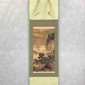 Op de afbeelding: Een traditionele Japanse hangrol met een landschapsschilderij in bruine en zwarte tinten. Het kunstwerk toont bergen, bomen en een klein gebouw, met een lichtgroene en olijfgroene rand en bovenkant. De rol wordt opgehangen door een zwarte staaf en touw.