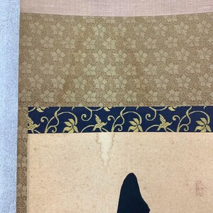 May include: Close-up of a traditional Japanese scroll. The scroll features a tan background with a black ink painting. The top section has a tan fabric border, followed by a gold floral patterned section, and a navy blue border with gold floral accents.